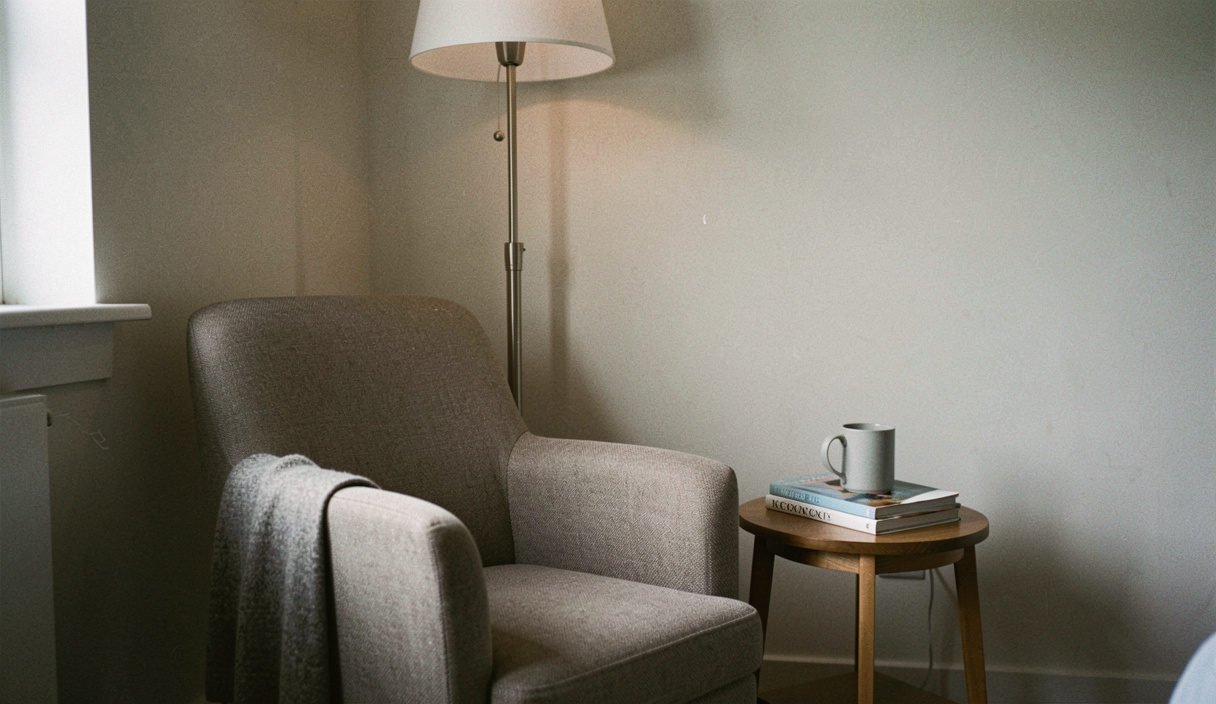 A low armchair in the corner of a room with a floor lamp behind it, a small side table holding a mug and a paperback, a plain wall to one side
