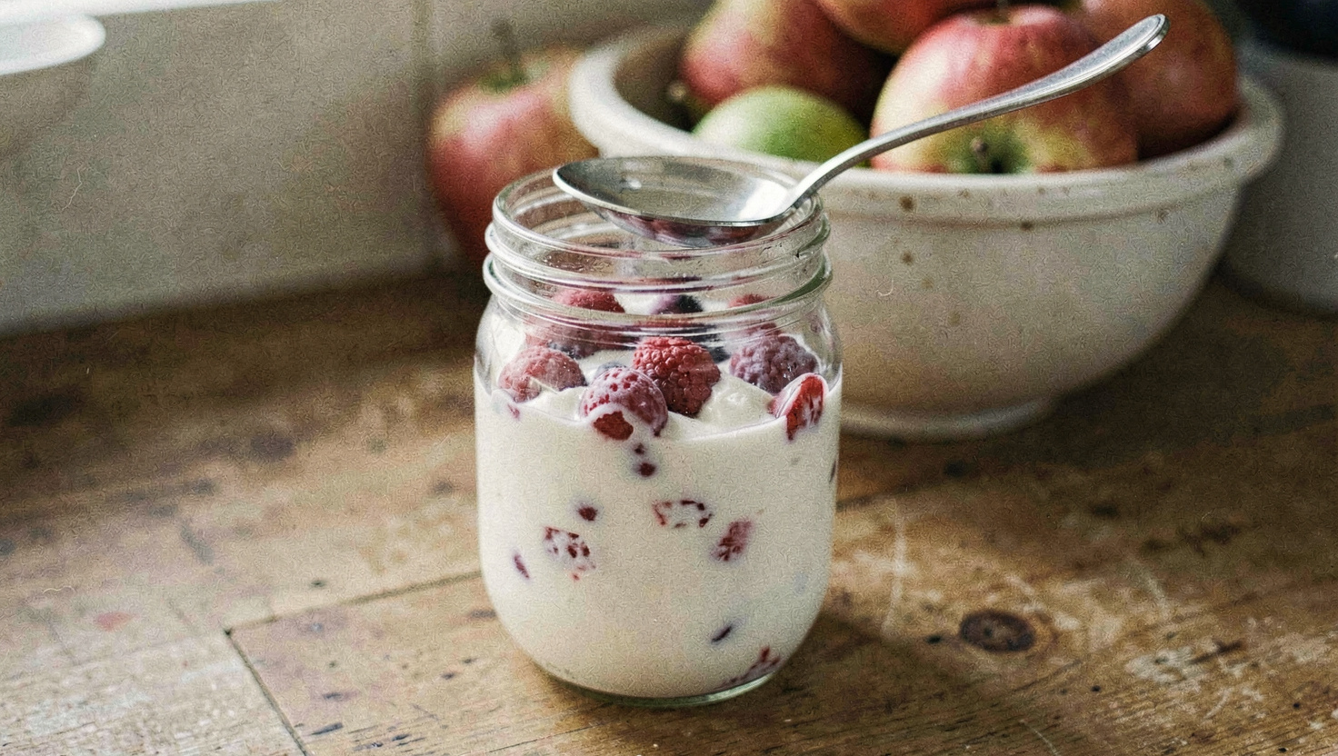 A glass jar of plain yoghurt with frozen berries stirred through it on a wooden counter, a bowl of apples behind and a spoon resting on the jar lid